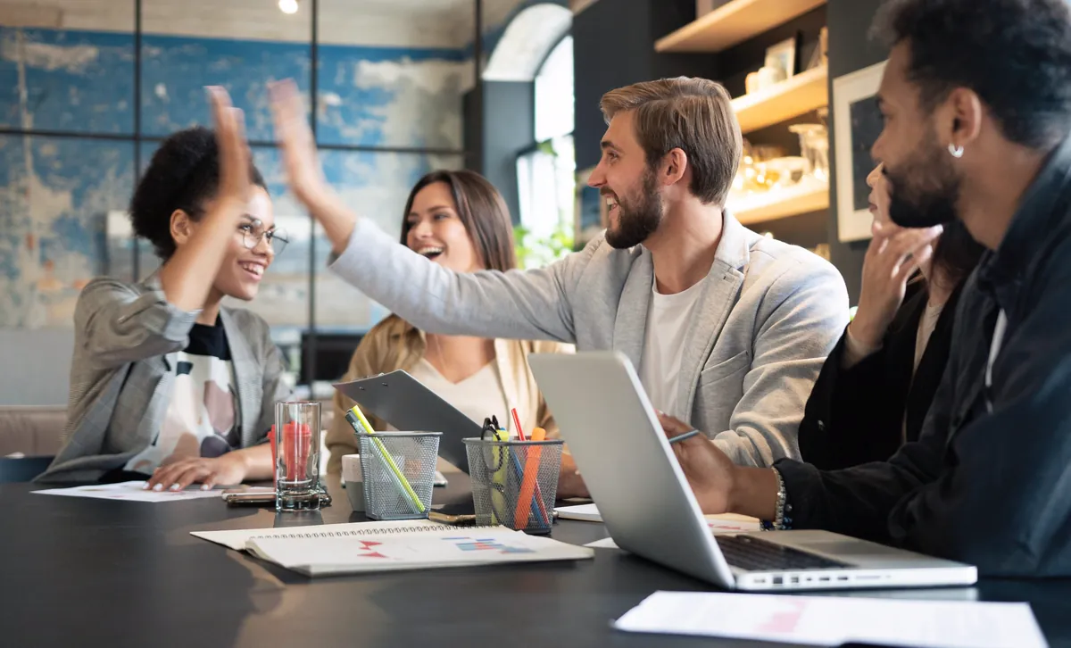 Smiling colleagues high-five in a meeting with a laptop and charts on the table, celebrating the team's productivity