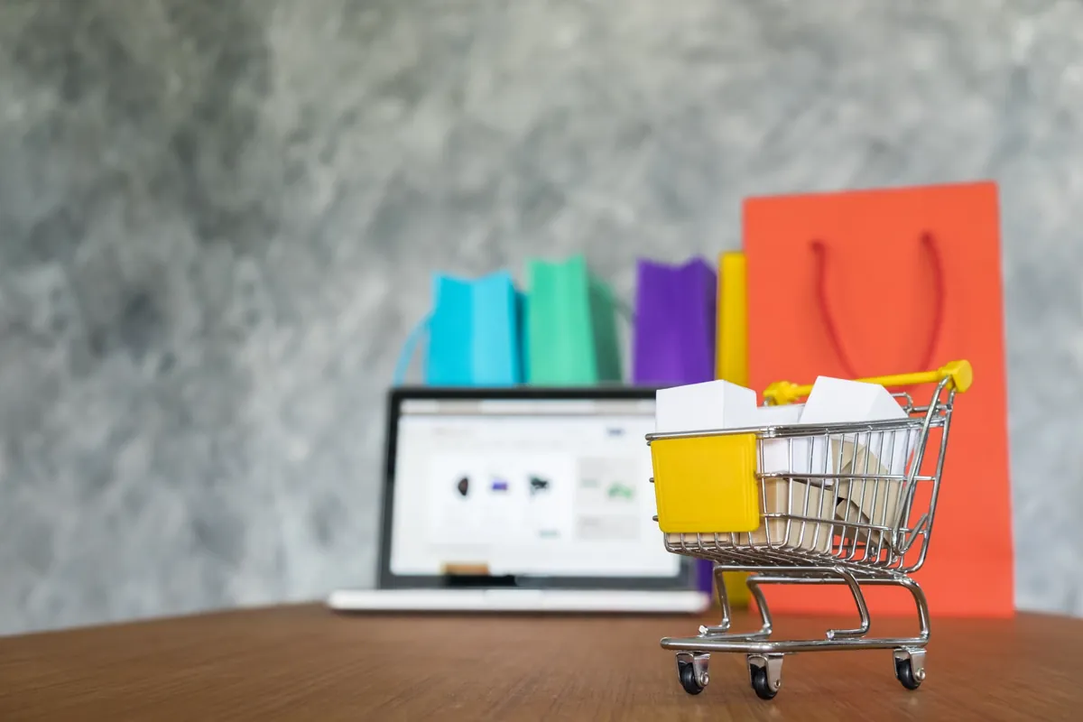 Miniature shopping cart with small boxes in front of a laptop showing an online store and colorful shopping bags, representing online sales