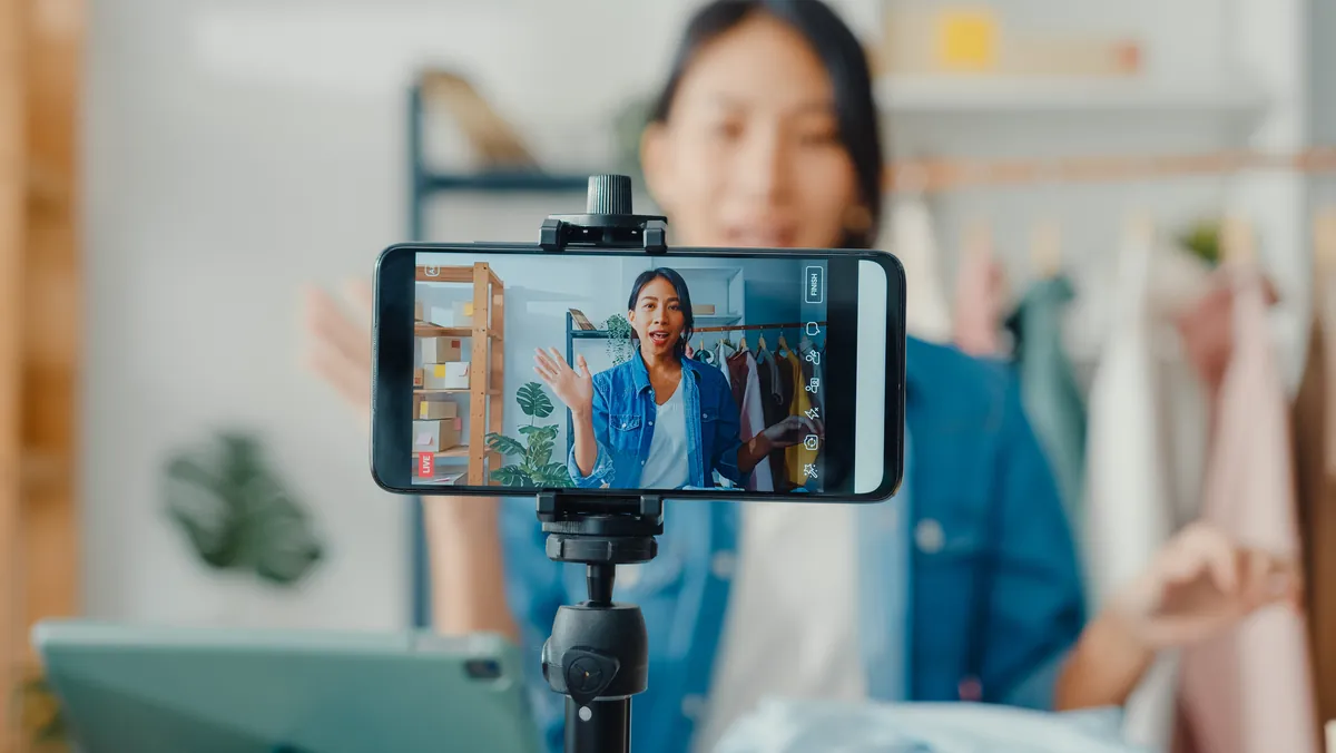 Phone on a tripod livestreaming a woman waving in front of a clothing rack and boxes, an example of digital entrepreneurship