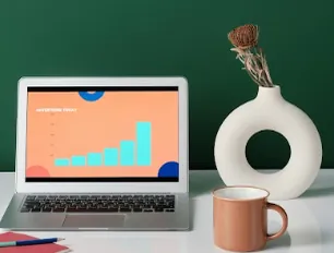 Laptop showing a rising bar chart on a white desk, next to a mug and a vase, suggesting marketing campaigns