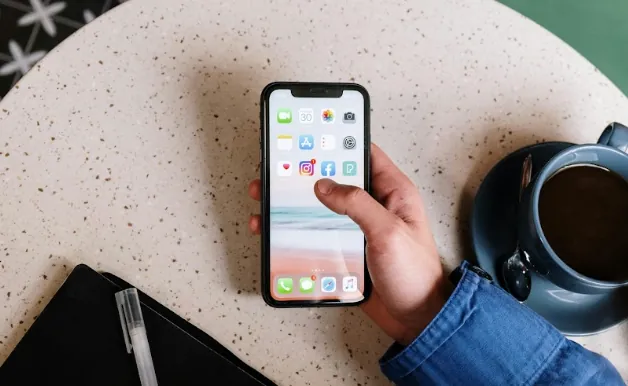 Hand holding a phone with the Instagram icon on the screen, over a terrazzo table with a cup of coffee and a notebook