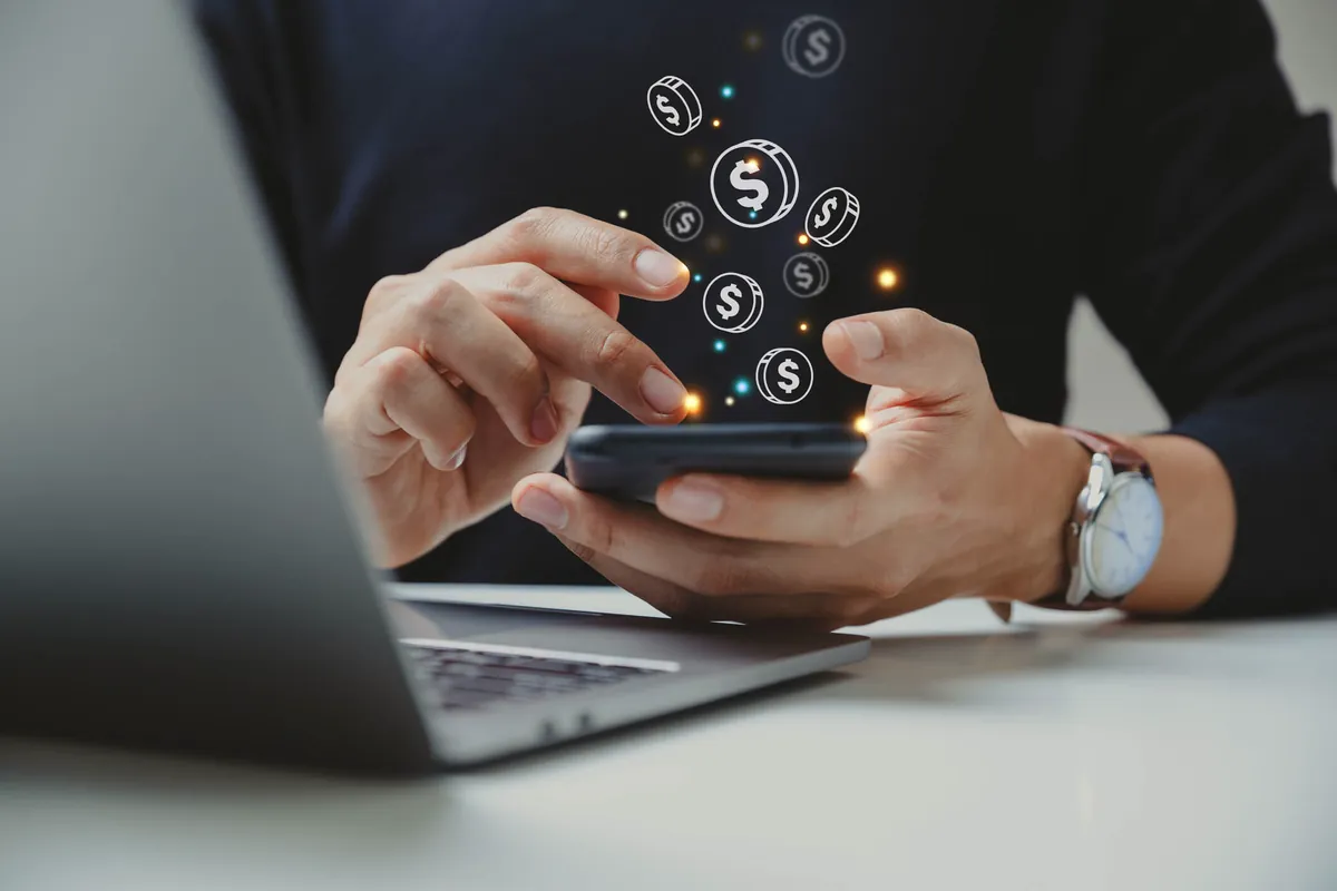 Hands using a phone next to a laptop with dollar coin icons coming out of the screen, representing making money online