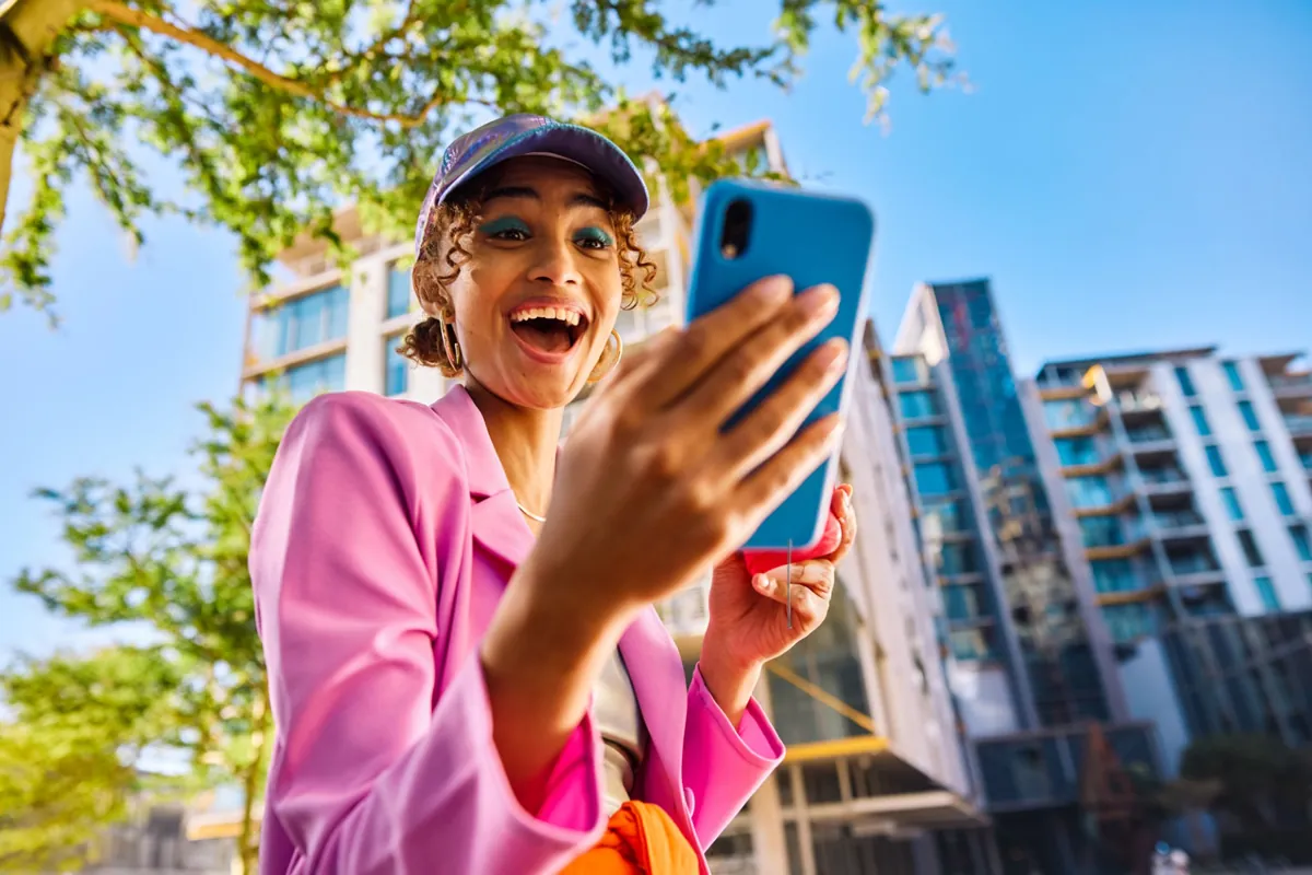 Young woman in a pink blazer and cap smiles excitedly at her phone on the street, a symbol of creative digital entrepreneurship