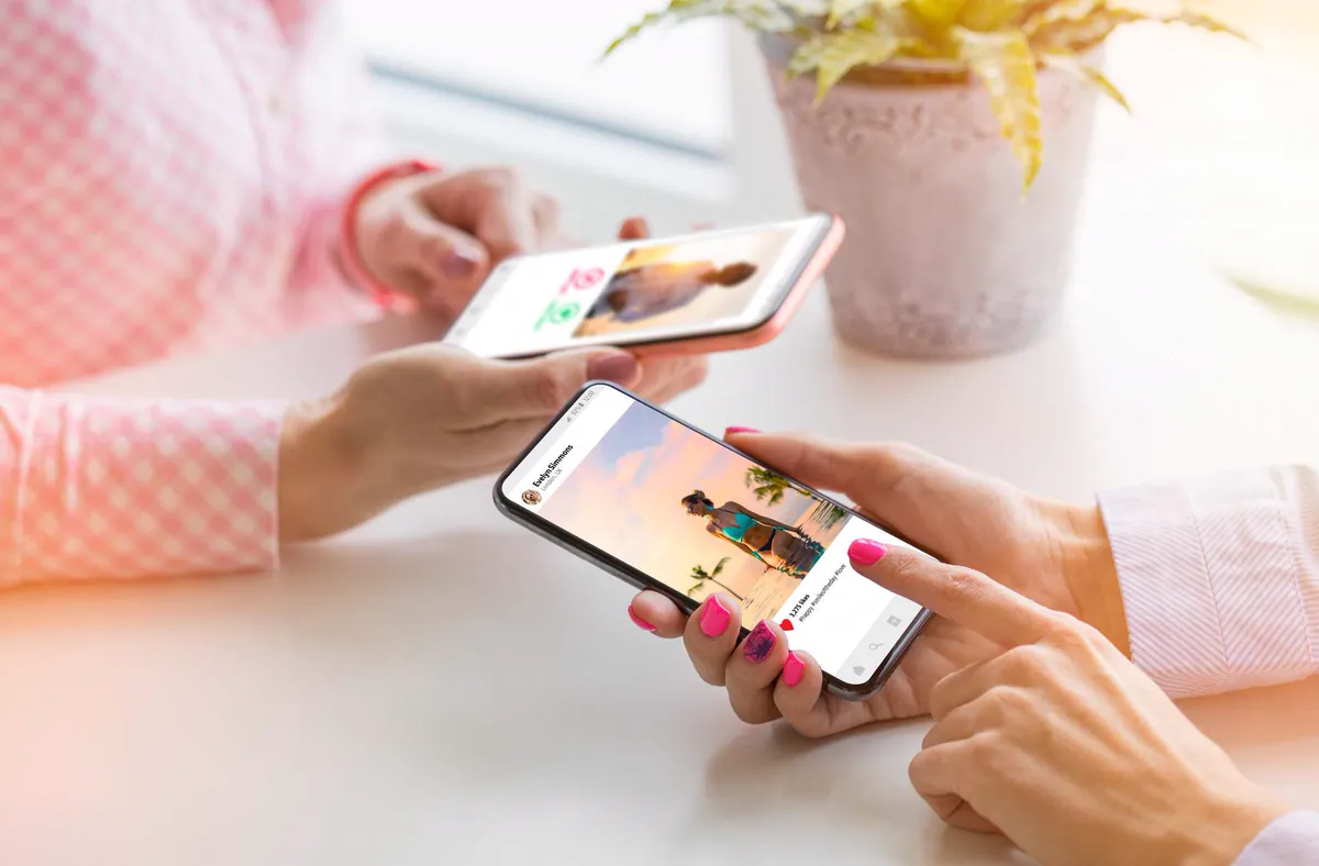 Two people using smartphones on a white table, one of them scrolling through a social media feed similar to Instagram