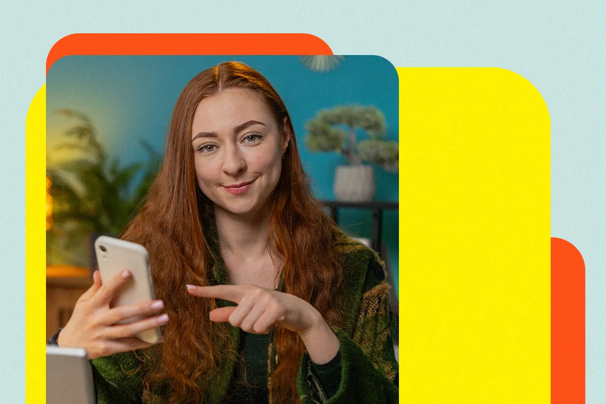 Mulher ruiva sorridente aponta para o celular na mão, sugerindo um link na bio que vende, sobre fundo amarelo e laranja