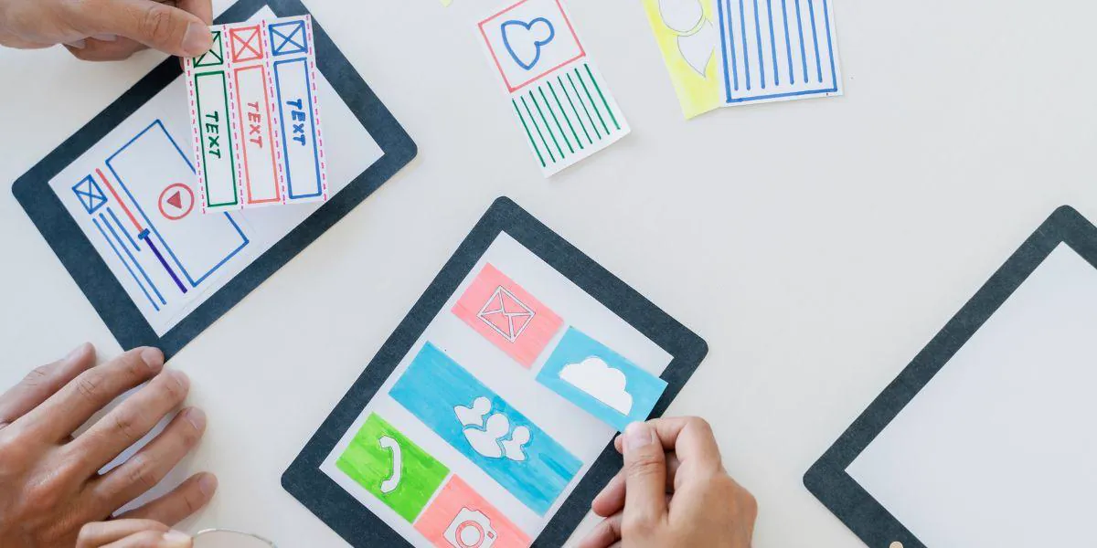Hands assembling paper prototypes with colorful icons on tablet frames, planning the user experience