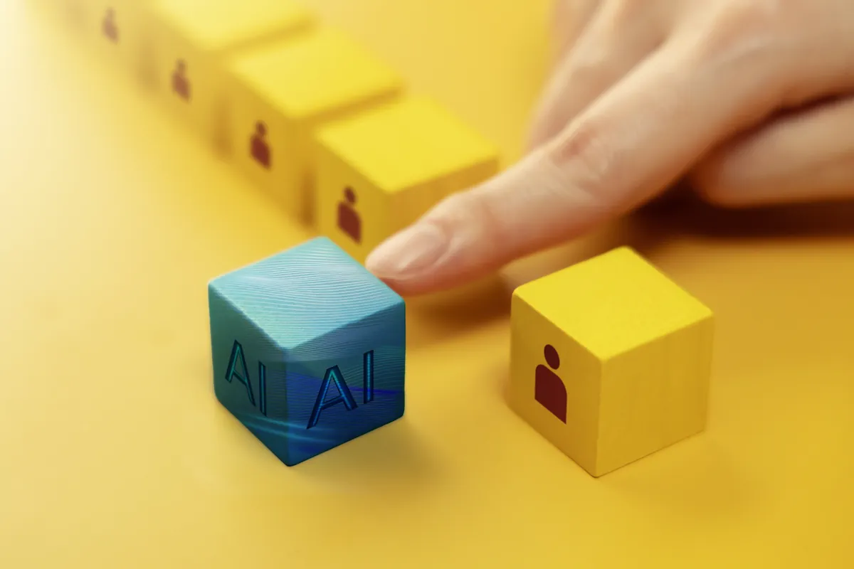 Finger pushes a blue cube with the letters AI toward a row of yellow cubes with people icons, suggesting the impact of AI