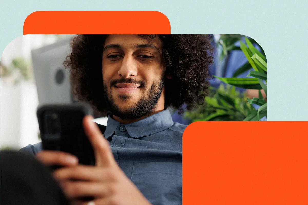 Man with curly hair and a beard smiles while looking at his phone, as if adjusting his Instagram bio, with plants in the background