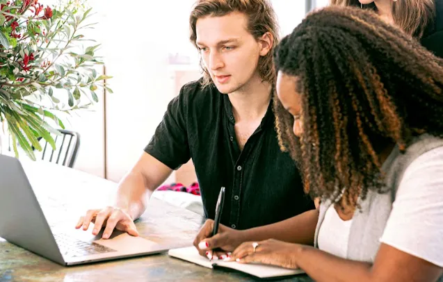 Man using a laptop and woman taking notes in a notebook at a table, planning how to promote affiliate links