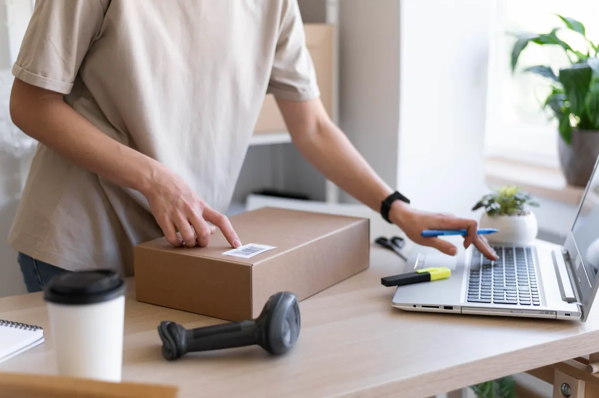 Person sticking a label on a cardboard box and typing on a laptop, preparing an order to sell more on Instagram