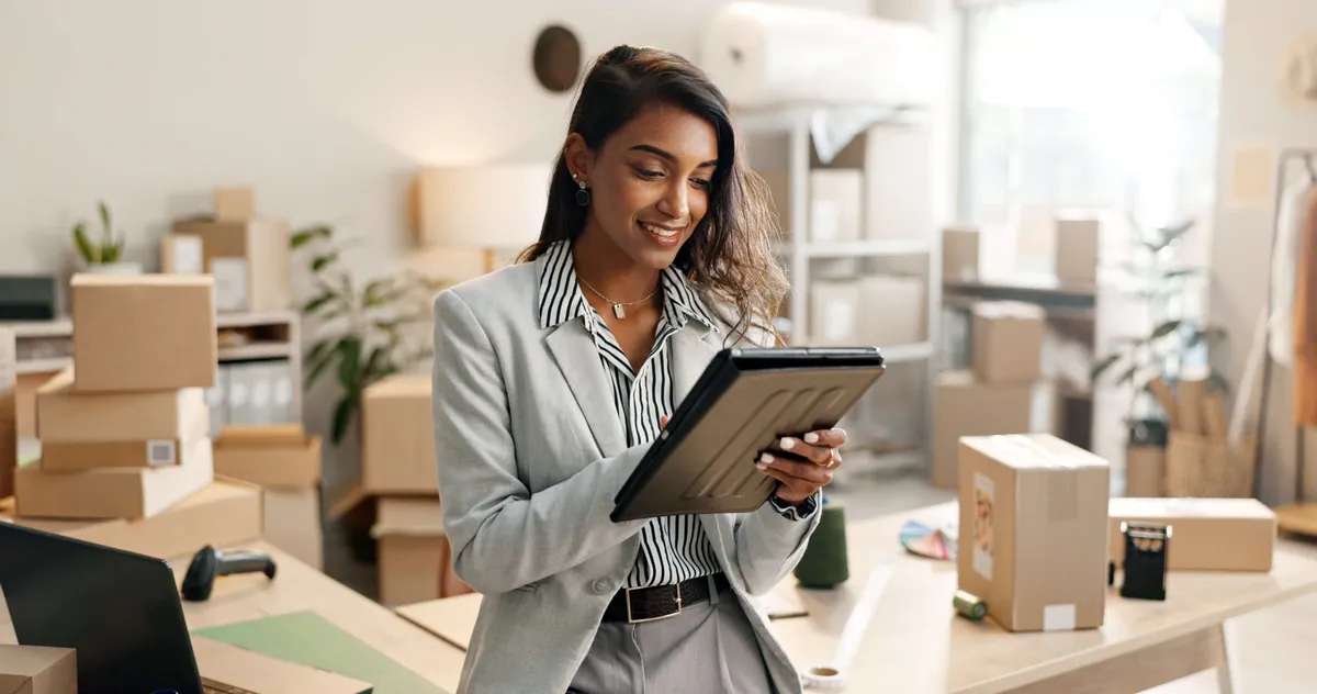 Smiling woman in a gray blazer using a tablet in an office full of shipping boxes, a typical day for someone who sells online