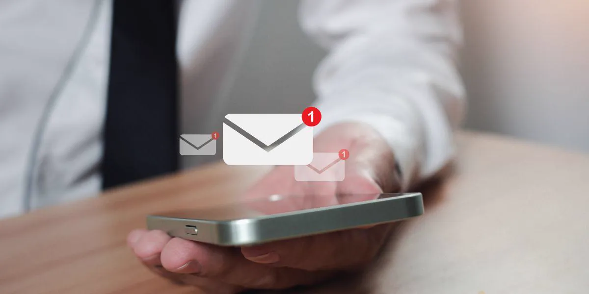 Person in a dress shirt and tie holding a phone with email and notification icons floating around, representing email marketing