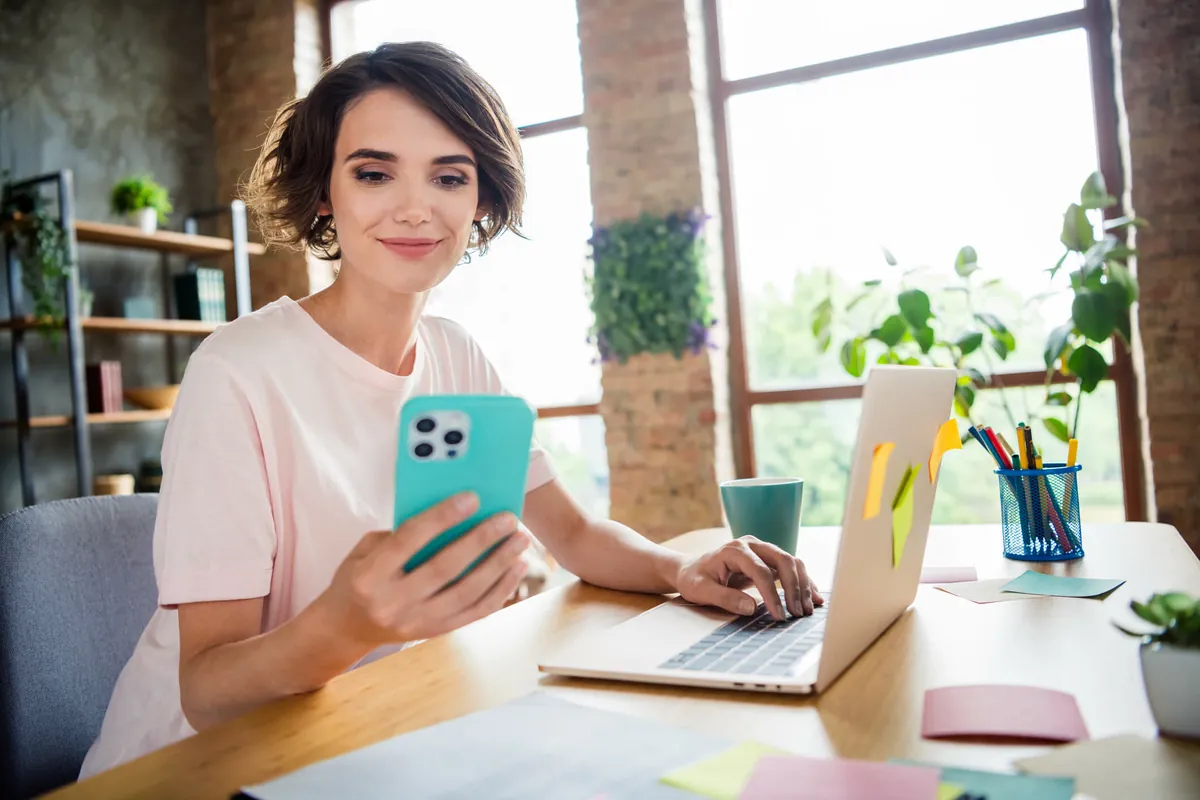 Smiling woman looks at a turquoise phone while typing on a laptop, as if adding a link to her Instagram bio