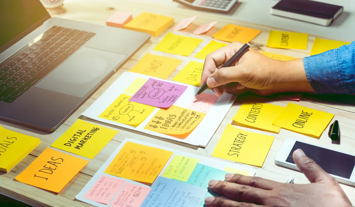 Hands writing on colorful sticky notes about digital marketing strategy, next to a laptop and a phone