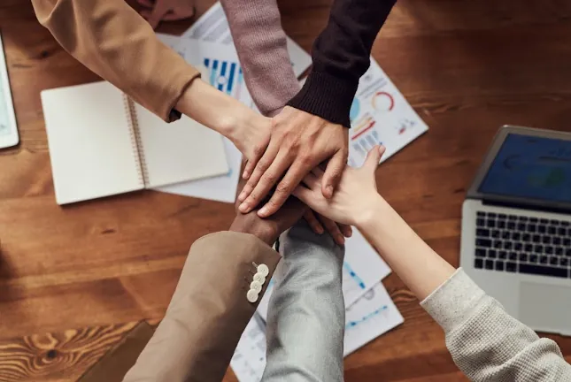 Five people's hands joined together over a table with charts, a notebook and a laptop, symbolizing partnerships and engagement